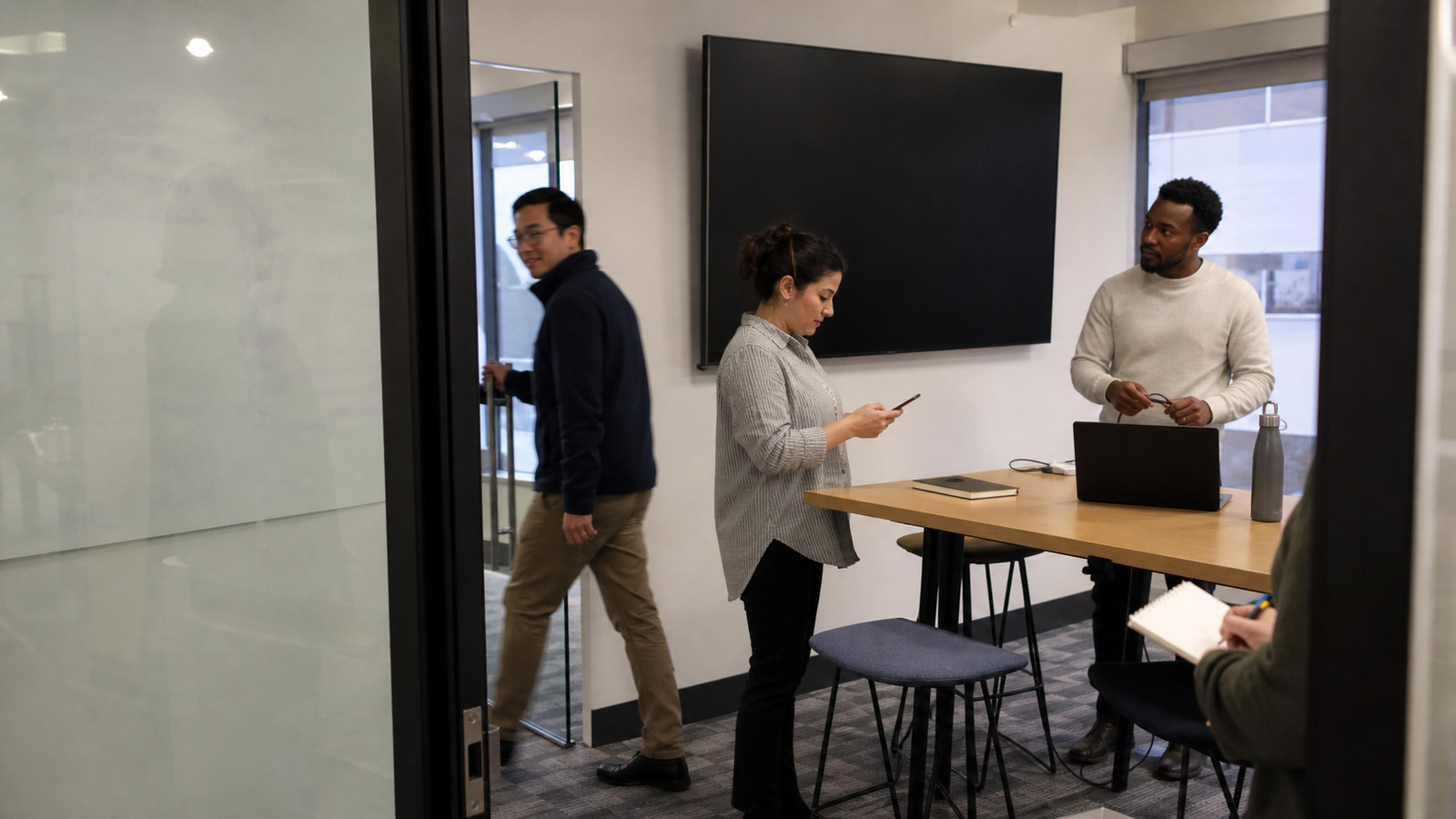 A small group around a high table in a meeting room, one person arriving through the door, screen dark on the wall.