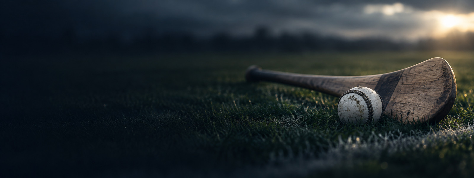 A hurley and sliotar lying together on cut grass, evening sun coming through low over the field.